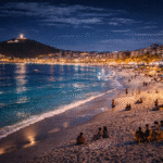 Praia do Forte em Cabo Frio à noite com areia branca iluminada, mar azul calmo e quiosques movimentados na orla com vista para o morro do Forte São Mateus.